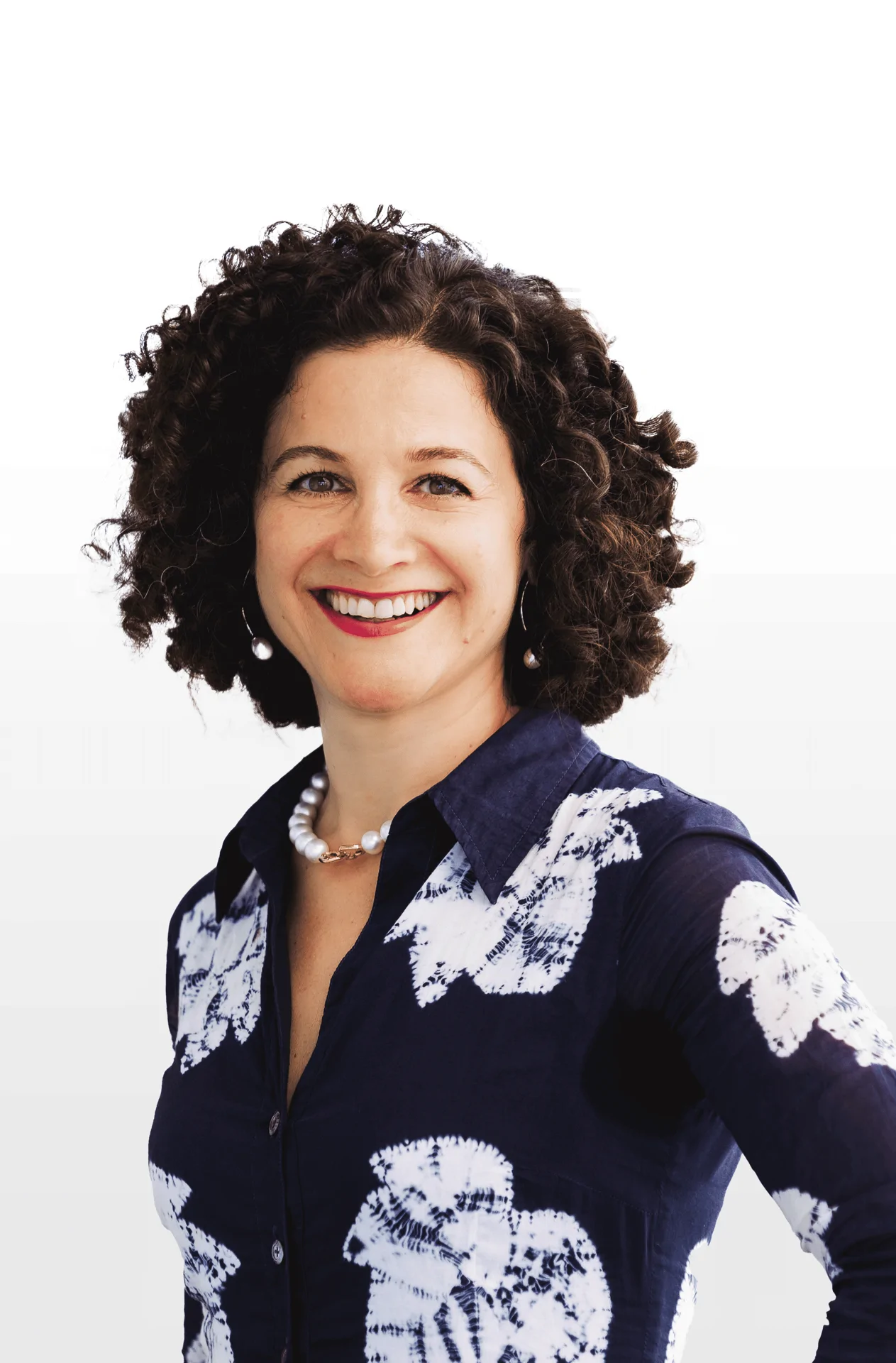 Woman with curly hair, wearing a navy shirt with white patterns and a pearl necklace, smiling at the camera.