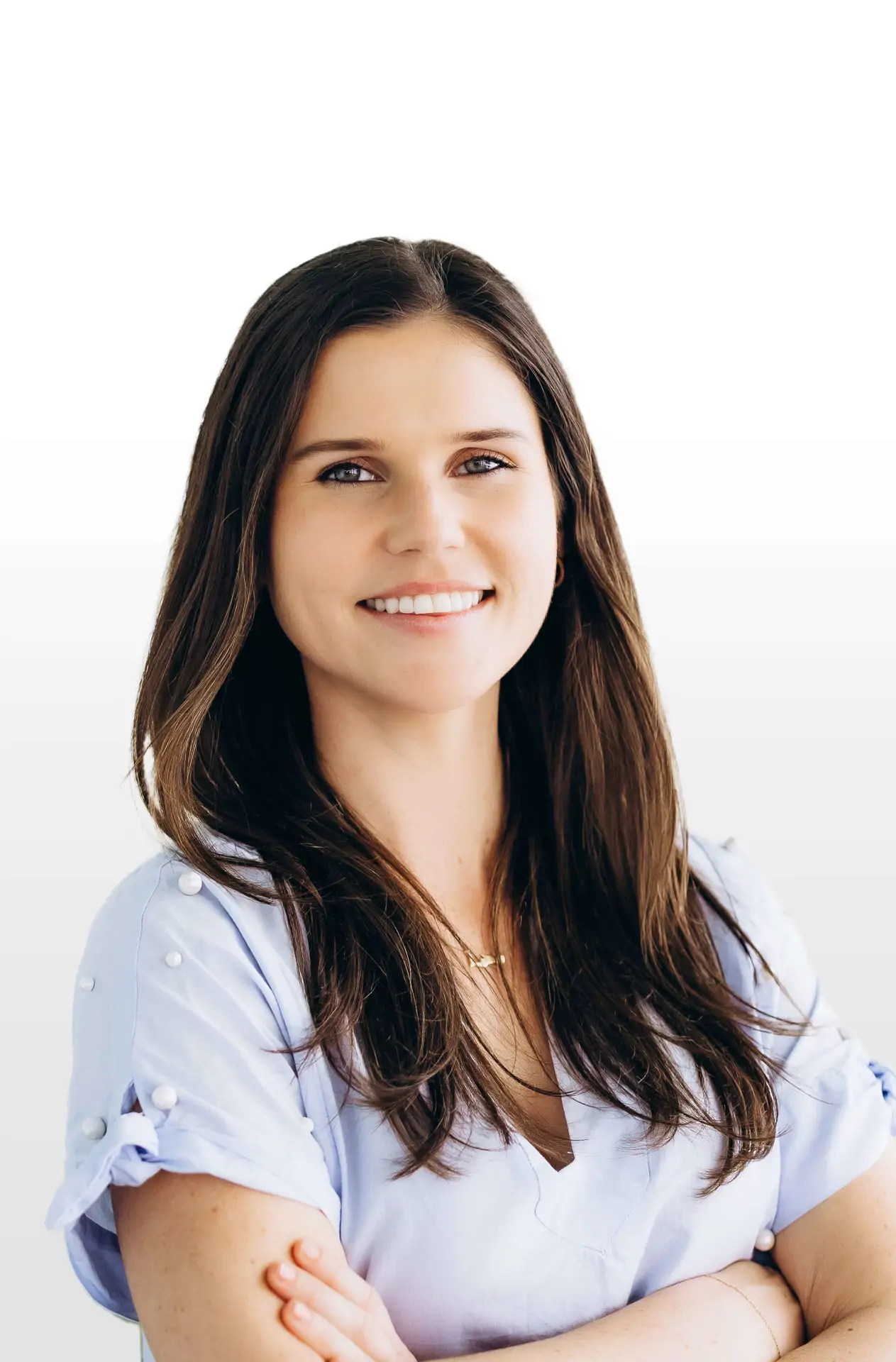 Smiling woman with long brown hair in a light blue blouse, standing with arms crossed against a white background.
