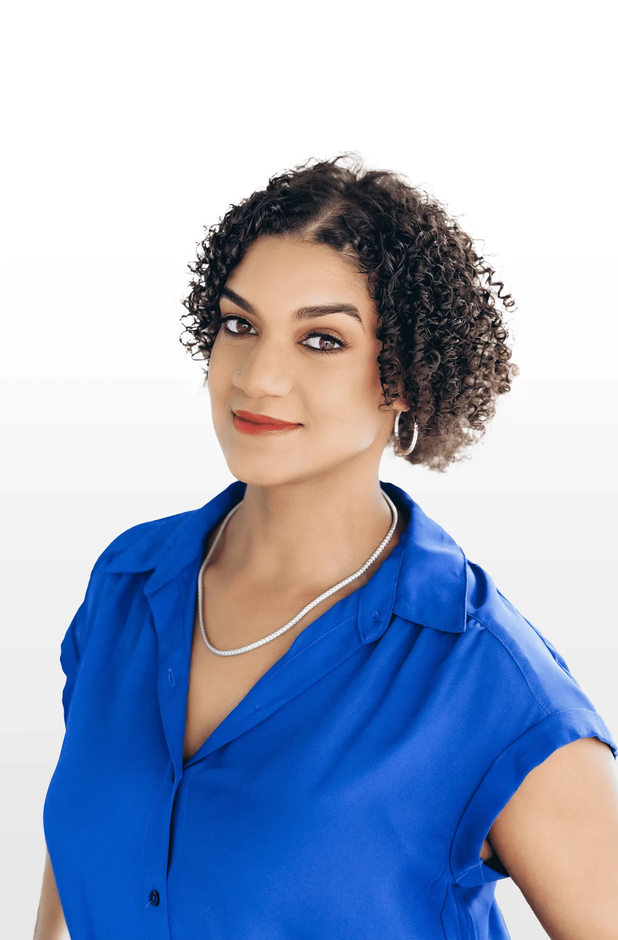 Woman with short curly hair wearing a blue blouse, silver necklace, and hoop earrings, smiling slightly at the camera.